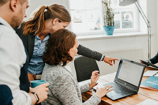 Eine Mitarbeiterin sitzt am Laptop und hinter ihr steht das Team mit Blick zum Laptop